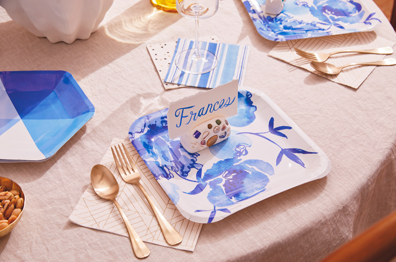 A party table set with plates, napkins and utensils in shades of blue, ivory and gold sit on top of a linen table cloth; on top of a square plate with a blue floral design sits a DIY mosaic place card holder with a handpainted place card that reads, 