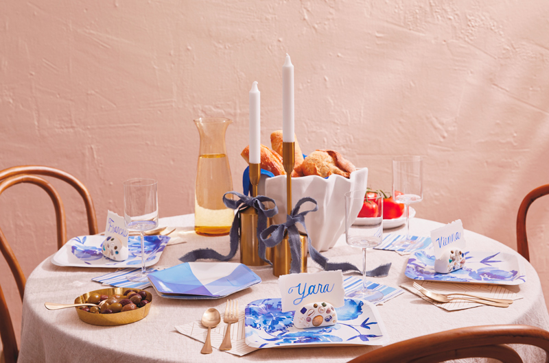 A party table is set with paper plates, napkins and utensils in shades of blue, ivory and gold; at the center of the table are gold candlesticks with white tapered candles in them; on top of each plate is a DIY mosaic place card holder with a handpainted place card.