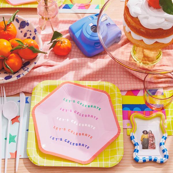 A party table set with plates and napkins in cheerful, bright shades of orange, yellow, pink, blue and green; next to the plate is a DIY personalized photo place holder that's been painted in bright colors and has a small Instax photo pasted inside the frame; a blue Instax camera sits at the center of the table between a bowl of mandarin oranges and a transparent yellow glass cake stand with a cake piled high with meringue frosting and a mandarin orange on top.