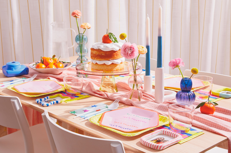 A party table is set with brightly colored plates, napkins and decor; each place setting is accompanied by a DIY photo frame personalized place card; at the center of the table is a transparent yellow glass cake stand holding a cake piled high with white meringue frosting and topped with a whole mandarin orange, still in its peel with the leaf attached; a bowl of more mandarins and a bright blue Instax camera sit on the table nearby.