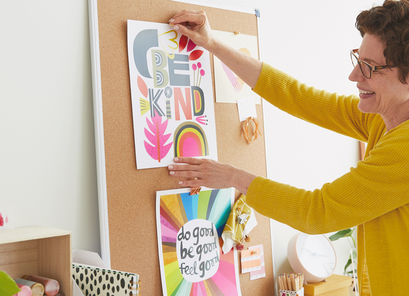 A woman smiles as she pins a free printable classroom poster to a corkboard; the poster is made in paper collage style and reads, 