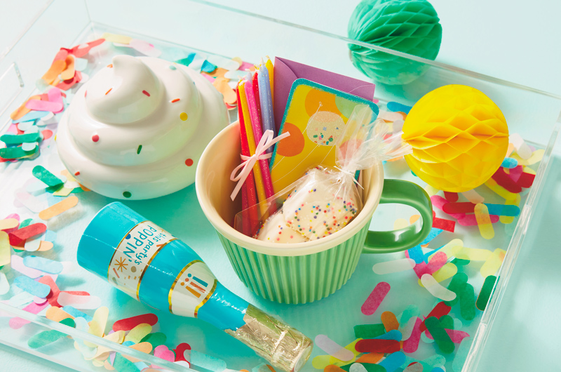 A light green mug that looks like a cupcake wrapper is filled with the makings of a small birthday celebration, including rainbow sprinkle-covered marshmallows, colorful glitter birthday candles, and a mini birthday card with balloons on the front; the mug is sitting in a clear acrylic tray with its lid, which is white and shaped like a swirl of cupcake icing covered in rainbow sprinkles; also on the tray is a party popper and colorful confetti.