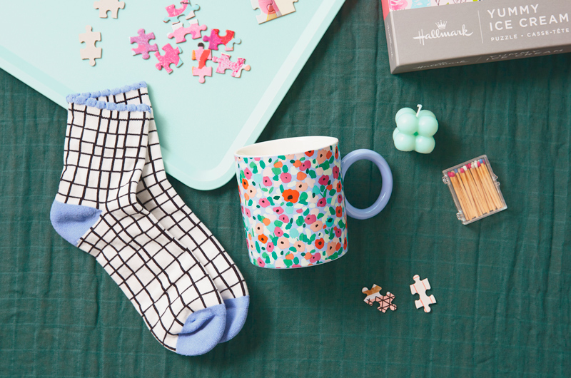 A ceramic mug covered in an impressionist-style floral pattern lays on a dark green blanket aside a pair of black and white grid pattern socks that have a light blue heel, toe and ankle trim; there is also a puzzle, a tea tray, a small candle and a tiny box of matches nearby, hinting at the idea of this being a cozy themed gift idea.