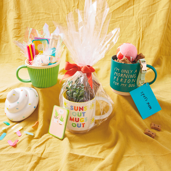A trio of mug-based gift bundles sit on a yellow fabric sweep; the three mug gift bundles are birthday, gardening, and pet themed.