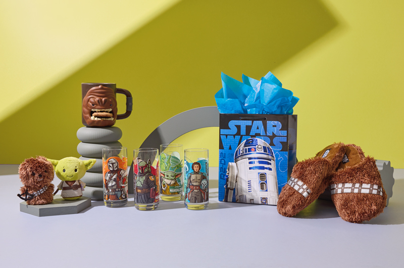 A selection of gifts for parents who love Star Wars, including a pair of fuzzy, brown Chewbacca slippers; a gift bag featuring R2-D2; a set of drinking glasses featuring characters from the show The Mandalorian, including Grogu™ and Din Djarin™ on each glass plus Ahsoka Tano™, Boba Fett™, Bo-Katan Kryze™ and Fennec Shand™; Chewbacca and Yoda itty bittys plush; and a Rancor cookie holder mug.