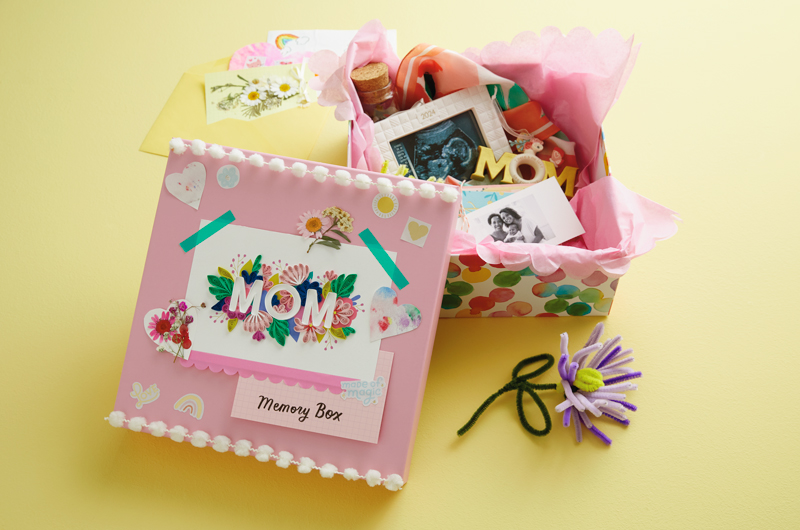A Mother's Day memory box with a light pink lid that's been decorated with a Mother's Day card and white pom-pom trim sits on a light yellow surface; inside the memory box are a Keepsake Ornament that reads 