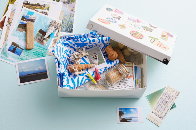 A memory box filled with items from various vacations and adventures sits on a light blue surface; the memory box is white, and the lid is covered in travel-themed stickers representing different countries; inside the box are concert ticket stubs, seashells, a small, swing-top jar with sand in it, drink umbrellas, a champagne cork, maps, postcards and a blue and white floral scarf.