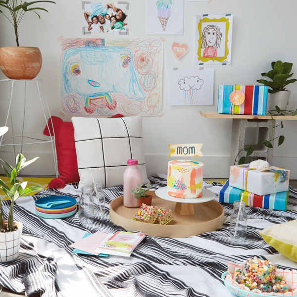 An indoor picnic setup for Mother's Day, with a black and white striped blanket on the floor; sitting on the blanket is a round wooden serving tray with a tall, round cake decorated with pale icing and a banner on top that reads, 