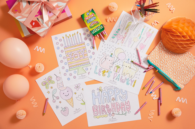 Printed coloring pages are colored in and scattered on a coral-colored surface; nearby is a tray full of crayons in different hues, gift boxes in shades of orange, white, pink and red, and light pink balloons.