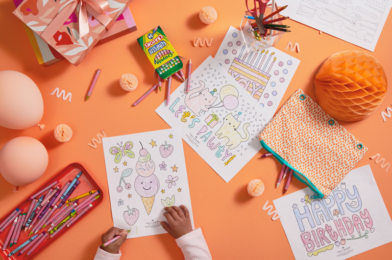 A little girl with a white bow in her hair colors in a free printable birthday coloring page on a coral-colored surface; surrounding her are more coloring pages, a tray full of crayons in different hues, gift boxes in shades of orange, white, pink and red, and light pink balloons.