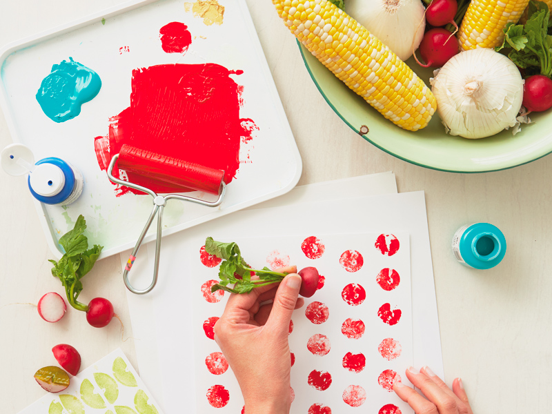 A woman uses a radish to create prints on a white piece of paper; the radish still has its leaves attached and has been cut on its horizontal axis, the interesting polka dot shapes it has left on the paper are a result of the red paint the vegetable was coated with before stamping.