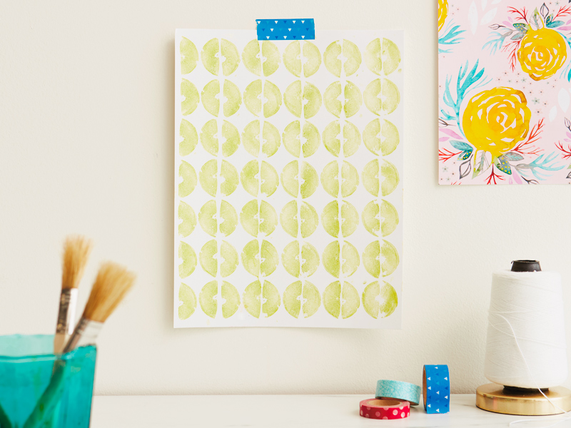 A vegetable stamping print hangs on a wall, secured by a piece of blue washi tape; the print is on a white piece of paper and consists of half moon shapes that are lime green in color, facing one another.
