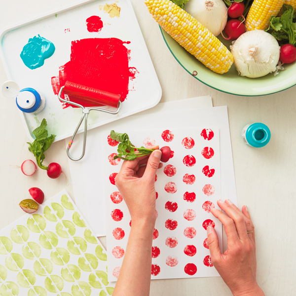 A woman uses a radish to create prints on a white piece of paper; the radish still has its leaves attached and has been cut on its horizontal axis, the interesting polka dot shapes it has left on the paper are a result of the red paint the vegetable was coated with before stamping.