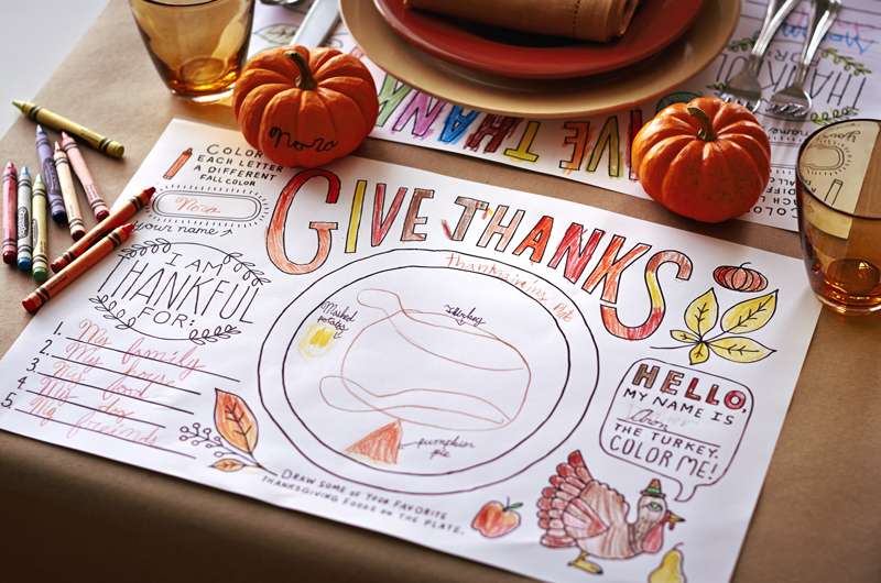 A free printable Thanksgiving activity in the form of a coloring placemat sits on a table surrounded by crayons, miniature pumpkins, and dinner plates.
