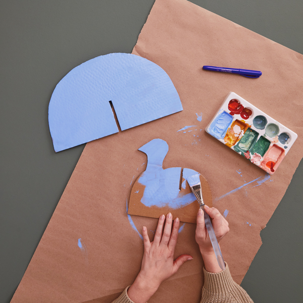 A woman's hands paint a piece of cardboard cut into a shape that resembles the profile of a turkey; nearby is a piece of cardboard shaped like a half circle with a notch cut into it; both pieces are painted a light blue.
