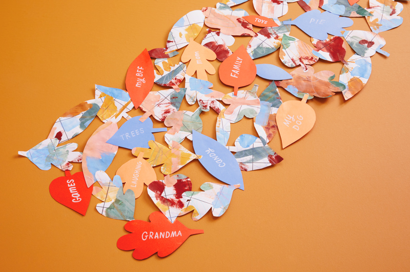 A gratitude table runner made with leaf-shaped cut-outs of wrapping paper and construction paper; written on the solid-colored pieces of construction paper are words like, 