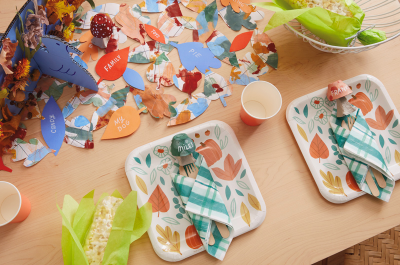 A Thanksgiving table set with fall-themed paper plates from the Hallmark Celebrate! partyware line; surrounding the plates are a variety of kids Thanksgiving crafts, like mushroom-shaped placeholders, popcorn 