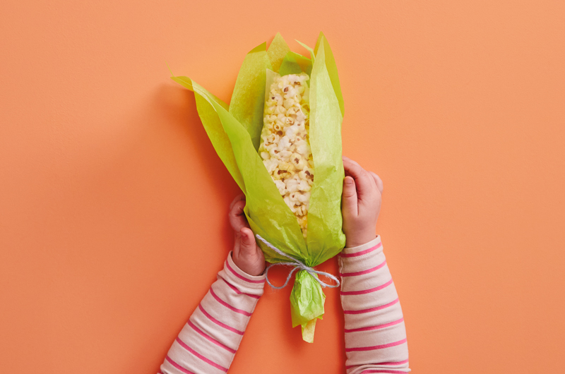 A child's hands reach into frame to hold a party favor shaped like a corn husk and filled with popcorn.