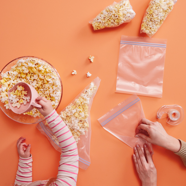 A child's hands scoops popcorn out of a bowl with a pink cup while a woman tapes back the bottom corners of a plastic storage baggie.