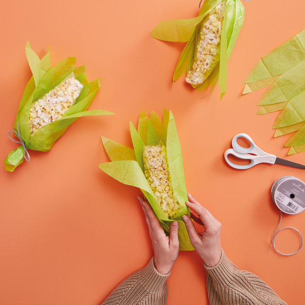 A woman's hands wrap pieces of green, cornhusk shaped tissue aper around plastic storage baggies filled with popcorn; nearby we see finished cornhusk popcorn party favors, a pair of scissors and a roll of twine.