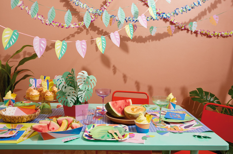 A table is set for a tropical theme party using various partyware products and table decorations, including a paper centerpiece made to look like a monstera plant in a pot, paper butterfly cupcake toppers, leaf-shaped paper plates, and brightly colored snack trays, cups and napkins; above the table hang three garlands strung with different shapes and colors of paper tropical leaves.
