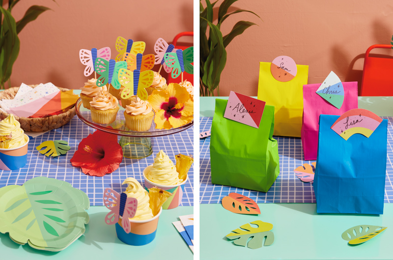 A cake stand set with half a dozen cupcakes with light orange frosting sits on a party table set for a tropical theme party; the cupcakes are topped with paper butterfly cupcake toppers and the table is strewn with leaf-shaped paper plates, brightly colored snack cups filled with pineapple dole whip, and brightly colored napkins. Beside this is a group of goodie bags in solid neon colors like blue, lime green, hot pink and bright yellow, with placecards repurposed as bag tags taped to the top.