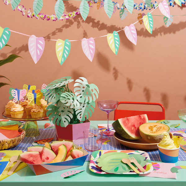 A table is set for a tropical theme party using various partyware products and table decorations, including a paper centerpiece made to look like a monstera plant in a pot, paper butterfly cupcake toppers, leaf-shaped paper plates, and brightly colored snack trays, cups and napkins.