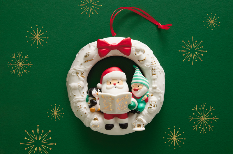 A Keepsake Ornament in the shape of a white wreath with gold foil embellishments; in the center of the wreath sits a little Santa, an elf, and a penguin; the three are looking at an issue of the Dream Book, which is also white with gold foil embellishments.