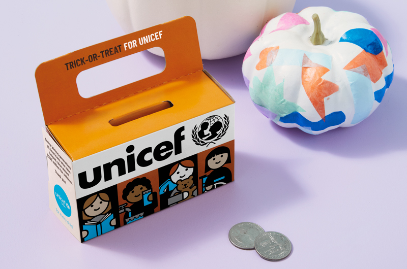 A Trick-or-Treat for Unicef box sits on a light purple surface along with a white mini pumpkin decoupaged with brightly colored tissue paper; sitting in front of the box is a pair of quarters—a seeming donation to the children Unicef supports.