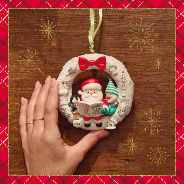 A Keepsake Ornament in the shape of a white wreath with gold foil embellishments; in the center of the wreath sits a little Santa, an elf, and a penguin; the three are looking at an issue of the Dream Book, which is also white with gold foil embellishments; the ornament sits on a wooden surface and a woman's hand is reaching into frame to pick it up.