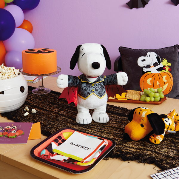 A table is set for a Snoopy-themed Halloween party; at the center of the table is an interactive Snoopy plush dressed like a vampire; surrounding him are other Peanuts products, like a floppy Snoopy plush in an orange and black Halloween print, a black pillow with a patch on it depicting a Vampire Snoopy and Vampire Woodstock popping out of a lit jack-o-lantern, and a papercraft card showing the entire Peanuts gang standing in a pumpkin patch on Halloween night.