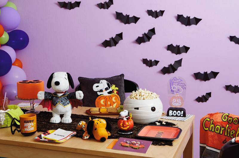 A table is set for a Snoopy-themed Halloween party; at the center of the table is an interactive Snoopy plush dressed like a vampire; surrounding him are other Peanuts Halloween products, including a plush Snoopy, a pillow, light-up decor, a tabletop figurine and a papercraft card; the table also is set with an orange-frosted cake that's been decorated to look like a jack-o-lantern, and a treat bowl filled with popcorn that looks like a round, wrapped mummy head; behind the table is a light purple wall decorated with black paper bats.