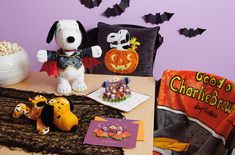 A table is set for a Snoopy-themed Halloween party; at the center of the table is an interactive Snoopy plush dressed like a vampire; surrounding him are other Peanuts products, like a floppy Snoopy plush in an orange and black Halloween print, a black pillow with a patch on it depicting a Vampire Snoopy and Vampire Woodstock popping out of a lit jack-o-lantern, and a papercraft card showing the entire Peanuts gang standing in a pumpkin patch on Halloween night.