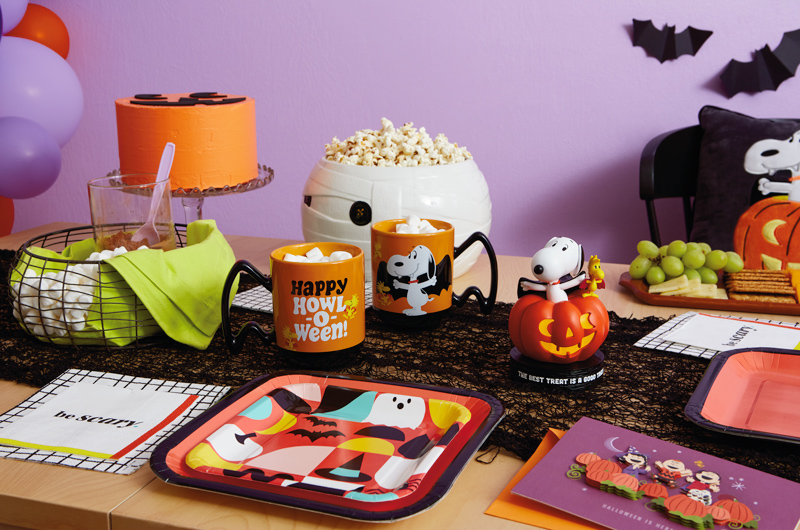 A table is set for a Snoopy-themed Halloween party; at the center of the table is an interactive Snoopy plush dressed like a vampire; surrounding him are other Peanuts products, like a floppy Snoopy plush in an orange and black Halloween print, a black pillow with a patch on it depicting a Vampire Snoopy and Vampire Woodstock popping out of a lit jack-o-lantern, and a papercraft card showing the entire Peanuts gang standing in a pumpkin patch on Halloween night.