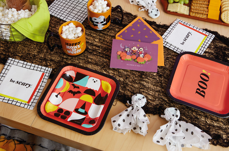 A table set for a Snoopy Halloween party features Snoopy mugs and a Peanuts paper craft card; there are also square paper party plates, one of which is orange with a black border and reads, 