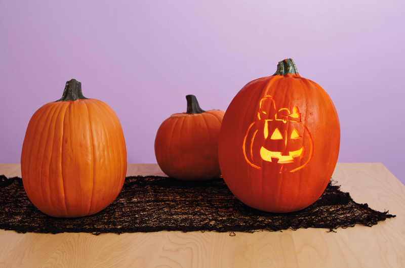 A pumpkin sits on top of a light wood table with a black mesh table runner, in front of a light purple walll; the pumpkin has been carved to feature a portrait of Peanuts Snoopy lying on top of a jack-o-lantern.