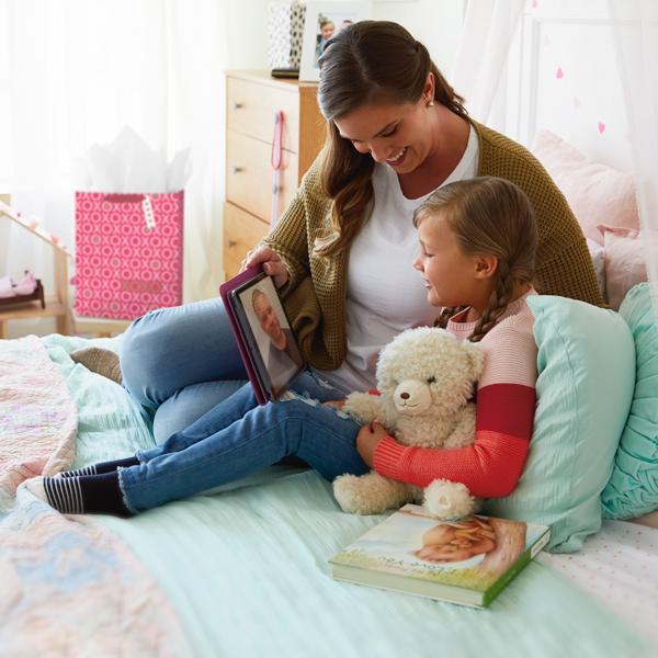A mother and her little girl sit on the little girl's bed as they chat with her dad on an iPad; he is in a military uniform; the little girl is holding a recordable plush bear and the coordinating recordable storybook sits on the bed next to her.