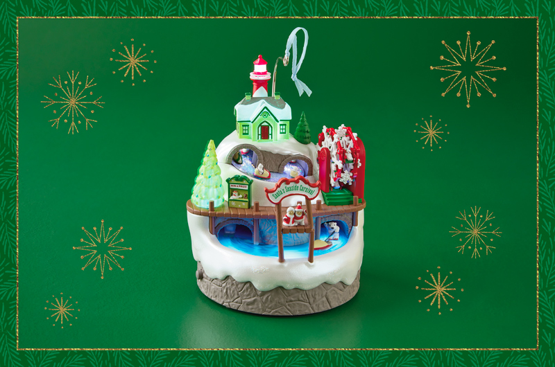 A Keepsake Ornament depicting a scene of a Seaside Carnival with a Christmas theme; Santa and Mrs. Claus wait at the end of a dock to greet newcomers; behind them is a snow-covered hill with a snowflake-shaped Ferris wheel, polar bears bobsledding through a series of light-up tunnels, and a light house on top that illuminates from the inside.