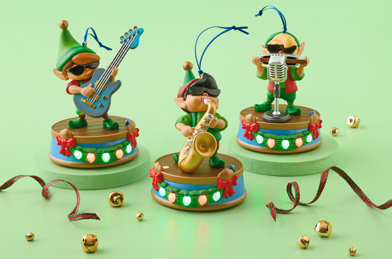 A trio of Keepsake Ornaments sits on a light green surface; each of the three ornaments is an elf in sunglasses, playing a jazz ornament; there is a blond elf playing a harmonica into a microphone, a black-haired elf playing a saxophone, and a brown-haired elf playing a guitar; the bases of each ornament light up and each ornament plays its own jazzy tune.
