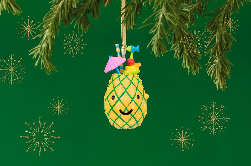A miniature Keepsake Ornament that looks like a pineapple, hollowed out to be used as a cocktail drink; sticking out the top of the pineapple is a drinking straw, a cocktail umbrella, and a cocktail toothpick garnished with tropical fruit and decorated on top with a fish.