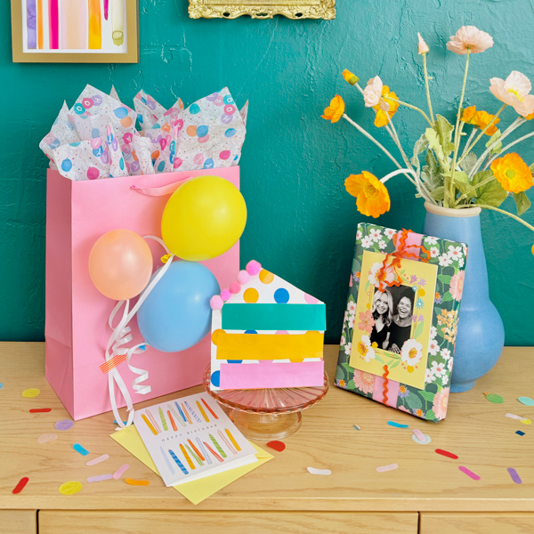 Three examples of unique, creative ways to wrap birthday gifts sit on a light-colored wooden table; one is a pouch decorated to look like a piece of birthday cake and filled with confetti and a gift card; another is a solid-colored gift bag with real balloons blown up to a small size and tied to ribbons, all taped to the side of the bag; the third is wrapped with floral gift wrap and is topped with a personalized photo card.