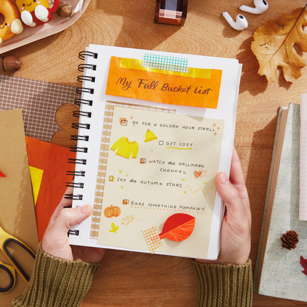 A woman's hands hold a folded back, spiral-bound notebook with a fall bucket list written on the page facing upward; the list is decorated with fall leaves cut out of orange, red and yellow construction paper.