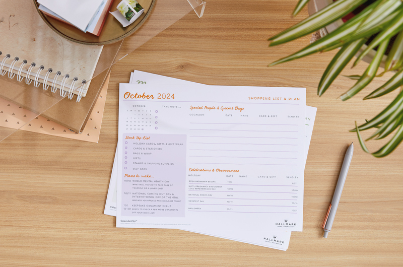 A printed version of the October 2024 planner page lays on a light wood desktop near a plant and a stack of journals and notebooks.