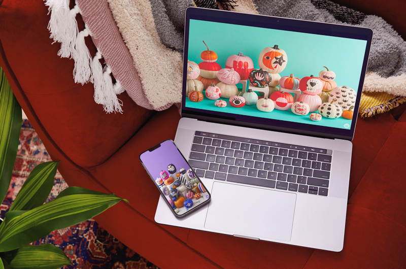 A laptop with a digital wallpaper displaying a collection of pumpkins painted for Halloween sits on a red couch; lying at the bottom of the laptop's keyboard panel is a smartphone displaying a mobile-sized digital wallpaper that shows a different collection of pumpkins painted for Halloween.