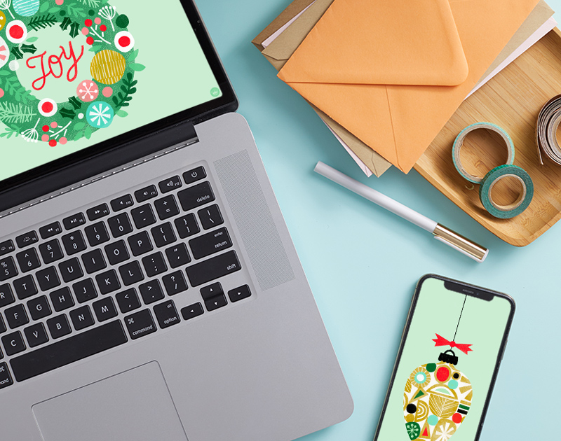 A laptop with a digital wallpaper featuring an illustration of a holiday wreath sits on a light blue desktop; nearby sits a smartphone displaying a different digital wallpaper that features an illustration of a holiday ornament.