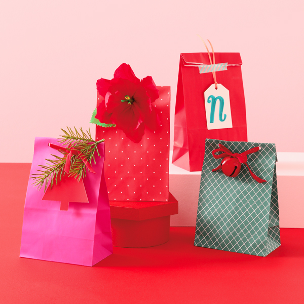 Four holiday goodie bags, each adorned with ribbon and an embellishment like a jingle bell, monogrammed tag, poinsettia flower and evergreen sprig, sit on a red tabletop.