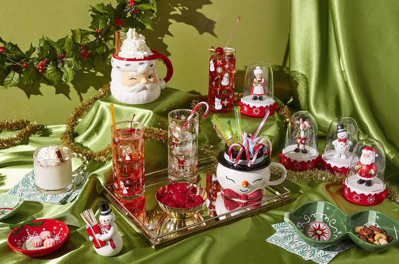 A table decorated with Christmas-themed glassware, mugs and serving dishes, with a shiny, green, satin tablecloth.