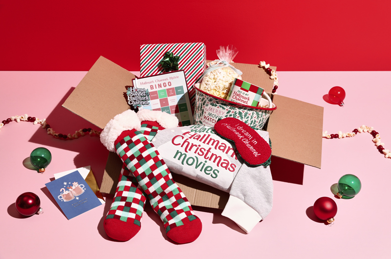 A Christmas care package meant for a Hallmark Channel lover that includes official Hallmark Channel gift product, such as a super-plush socks and eyemask set, a sweatshirt, an ornament, a Hallmark Channel Movie bingo card set, and a popcorn bowl with a mug and a bag of popcorn inside.