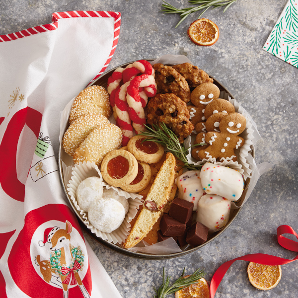 A decorative tin full of homemade Christmas cookies sits on a stone countertop next to a red and white holiday tea towel, some paper napkins covered in a holly pattern, dried orange segments, and rosemary sprigs.