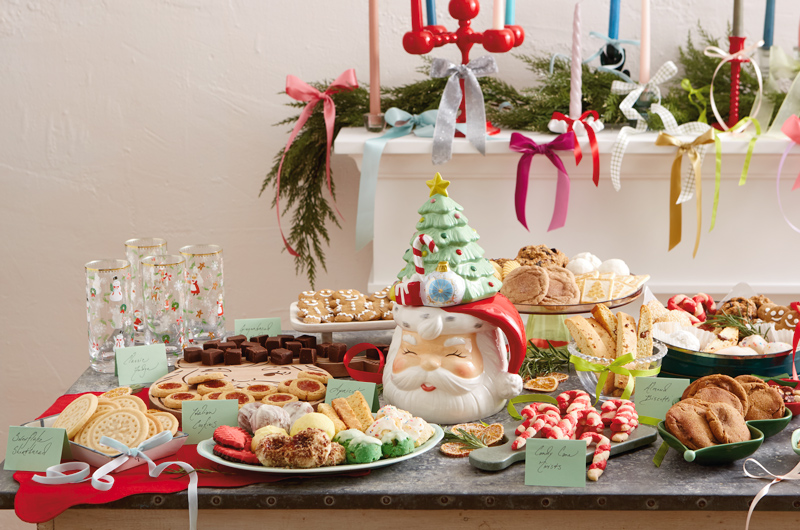 A stone countertop is covered with plates, bowls, serving dishes and trays that are filled with various homemade Christmas cookies; at the center of the cookies is a large, figural cookie jar in the shape of a smiling Santa; the lid of the cookie jar is a Christmas tree.